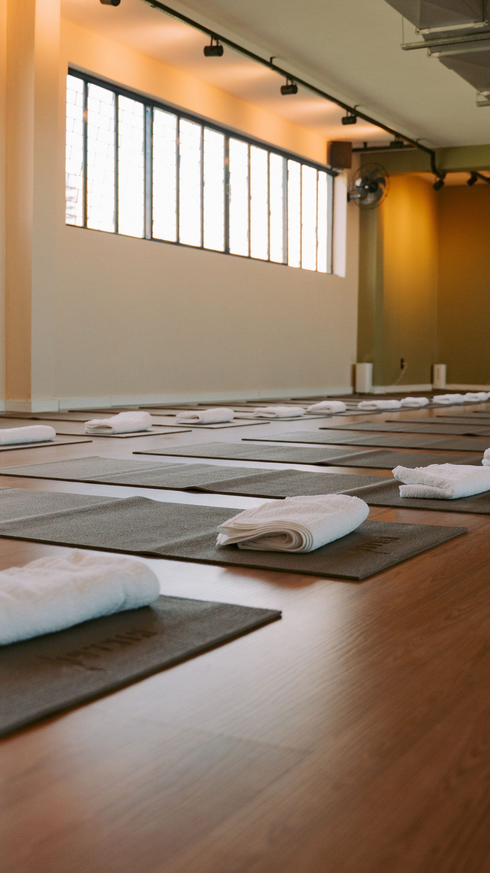 Sala de hot yoga do Hikari montada para a aula, com tapetes e toalhas sob a luz das janelas altas.