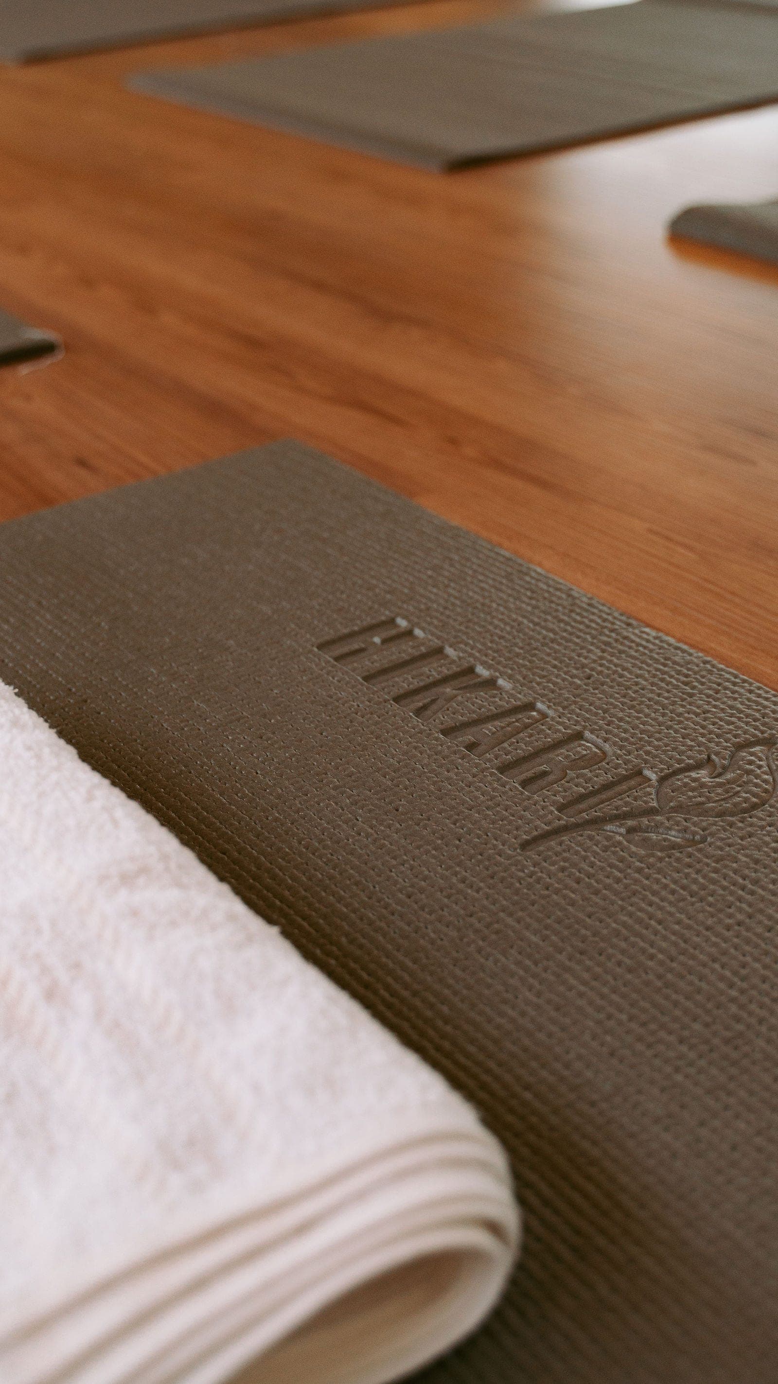 Tapete de yoga cinza com a marca HIKARI em relevo e uma toalha branca dobrada, sobre o piso de madeira do Studio Hikari.